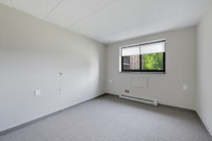 Interior Unit Bedroom, white walls, gray carpeting, large window on far-side wall.